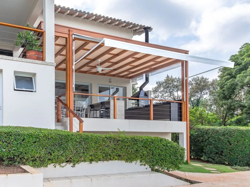 Terraço elevado de uma casa branca com um pergolado de madeira coberto por um toldo retrátil branco. A área possui uma churrasqueira e é cercada por uma sebe verde.