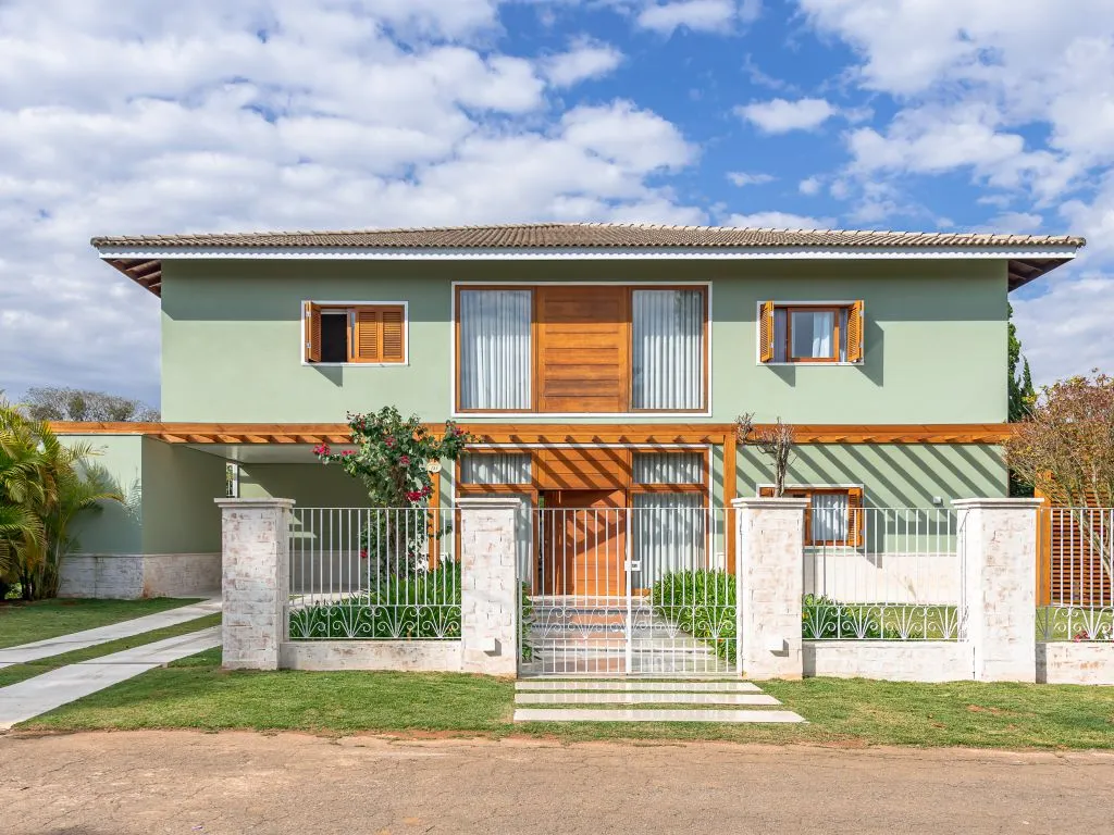 Fachada de um sobrado pintado de verde-claro, com janelas e porta de madeira, um muro branco com pilares de pedra e um pergolado de madeira sobre o portão de entrada.