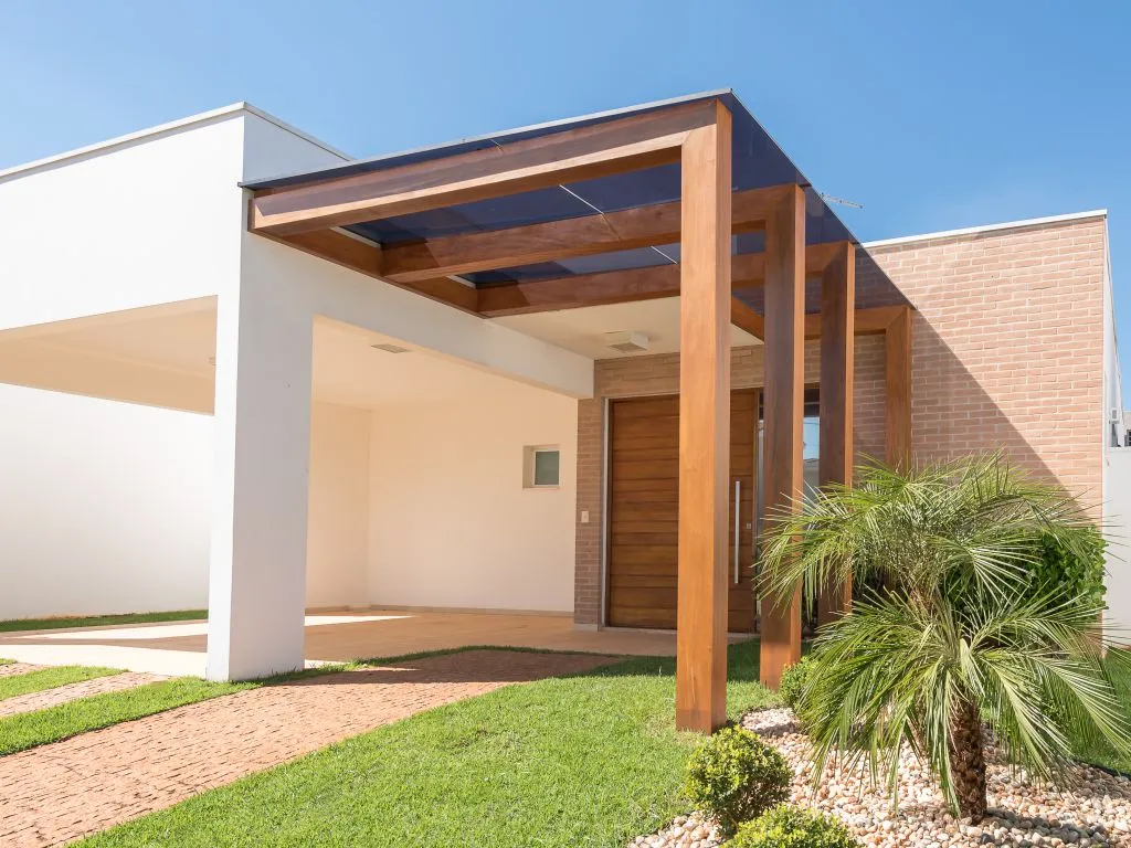 Entrada de uma casa térrea moderna com um pergolado de vigas de madeira e cobertura de vidro escuro sobre a garagem e a porta de entrada de madeira.