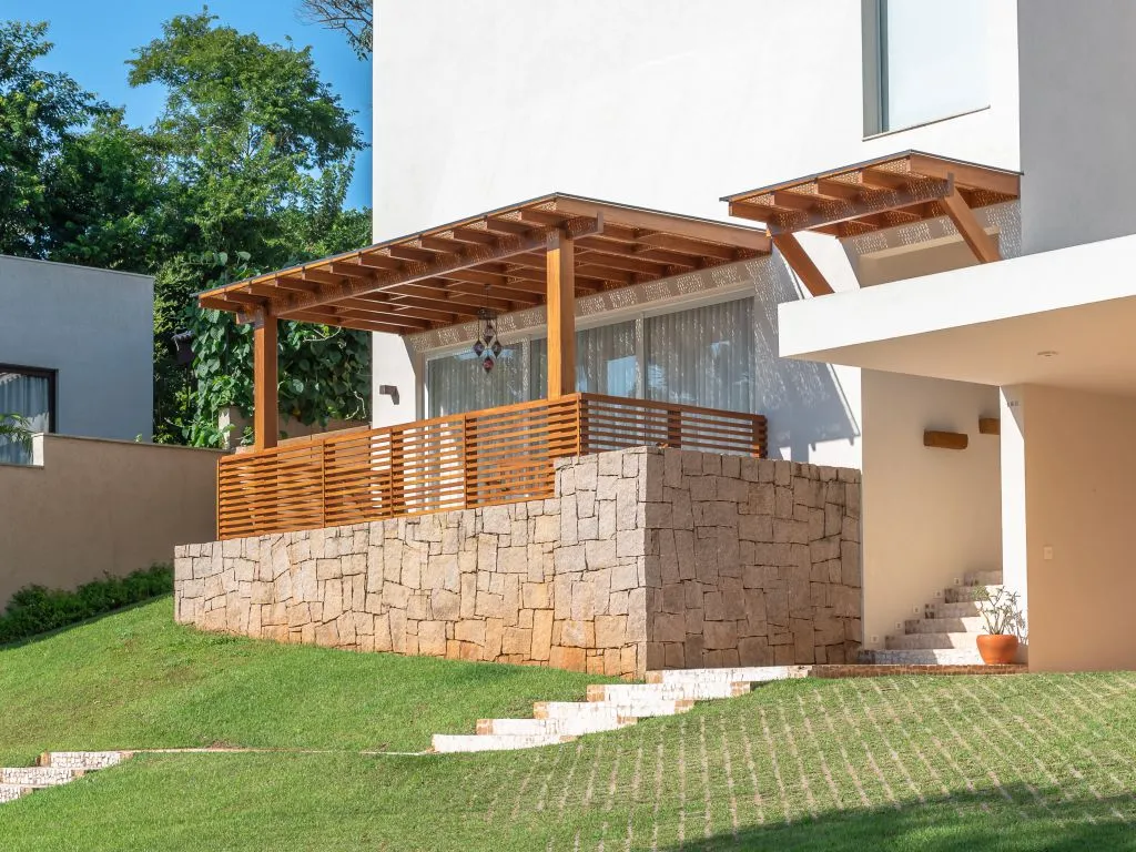 Casa moderna branca em um aclive gramado, com um terraço elevado sobre uma base de pedra, protegido por um guarda-corpo de ripas e um pergolado de madeira.