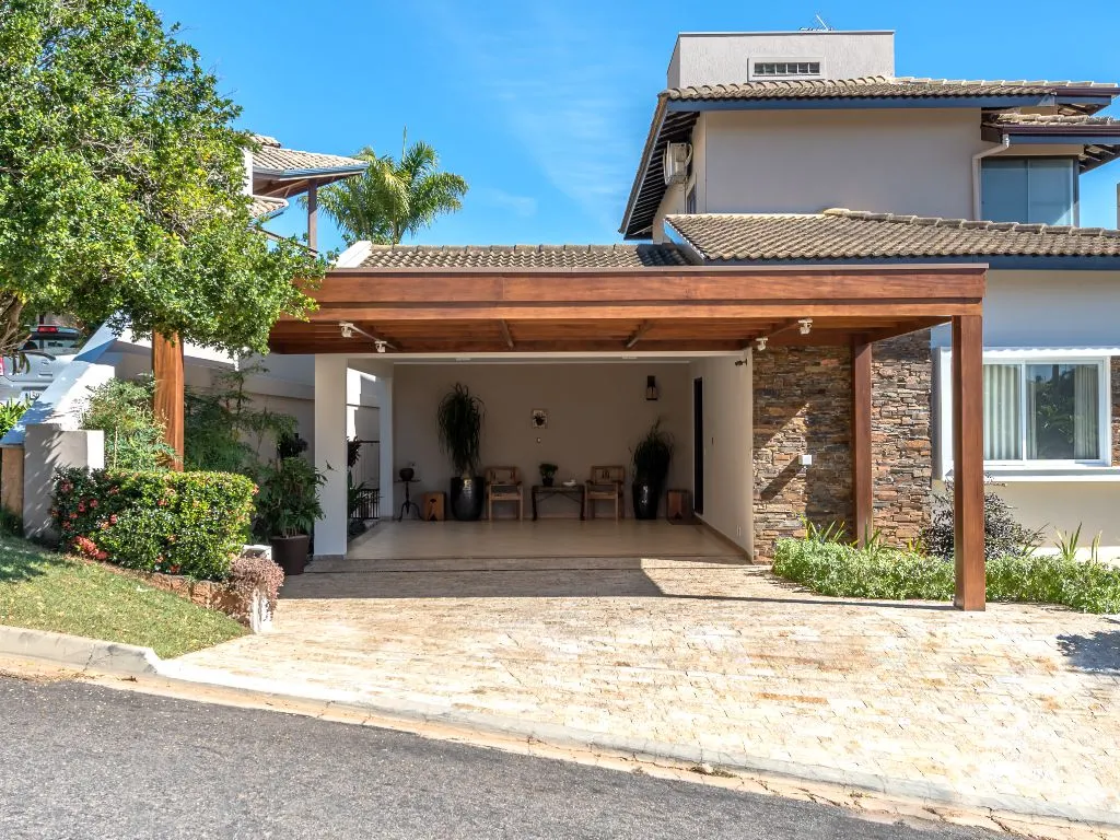 Fachada de casa moderna com um pergolado de madeira robusto cobrindo a garagem aberta, que tem uma parede de destaque em pedra e móveis ao fundo.