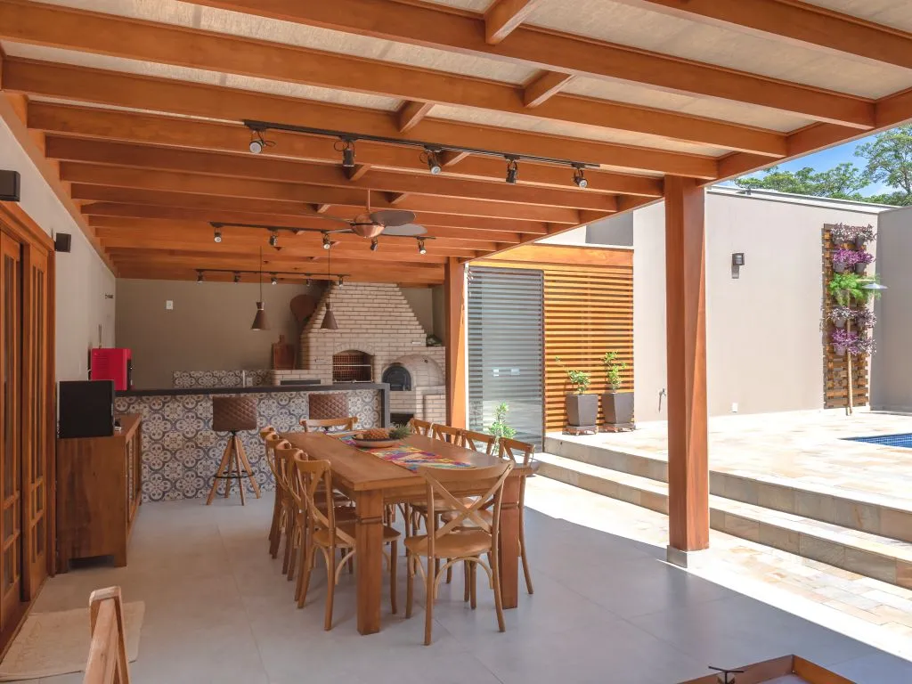 Pergolado de madeira com forro e ventilador, cobrindo área gourmet com churrasqueira de tijolos claros, balcão e mesa de jantar rústica