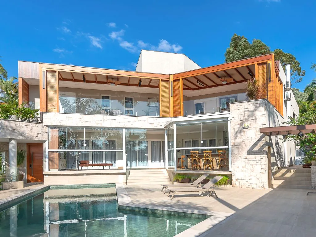 Casa moderna de luxo com piscina, apresentando varandas no andar superior cobertas por pergolados de madeira e uma fachada que combina pedra, vidro e madeira.