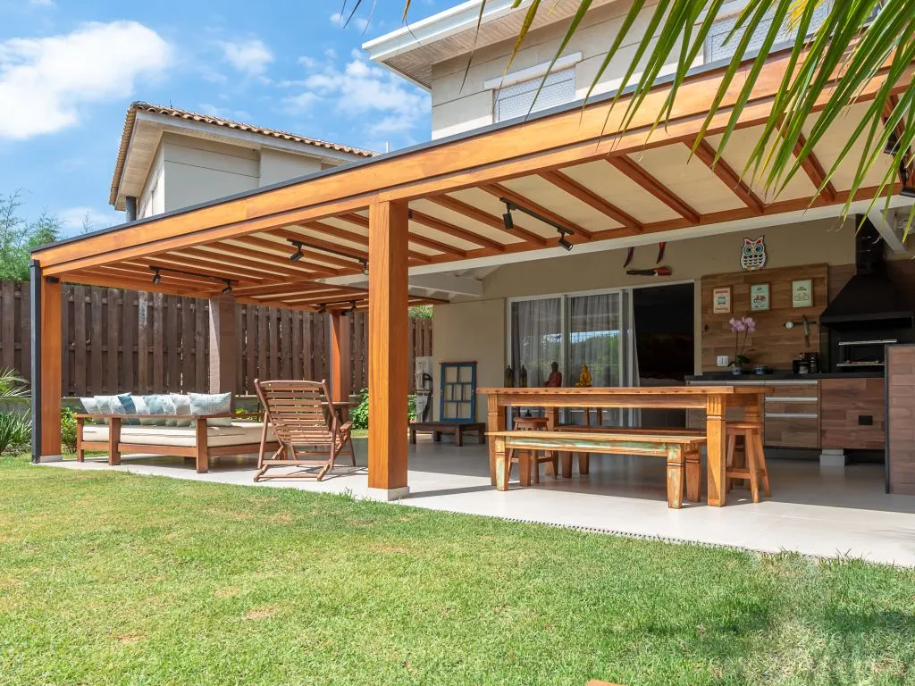 rea de lazer no quintal com um grande pergolado de madeira cobrindo um conjunto de mesa de jantar, bancos e um sofá, ao lado de um gramado verde.