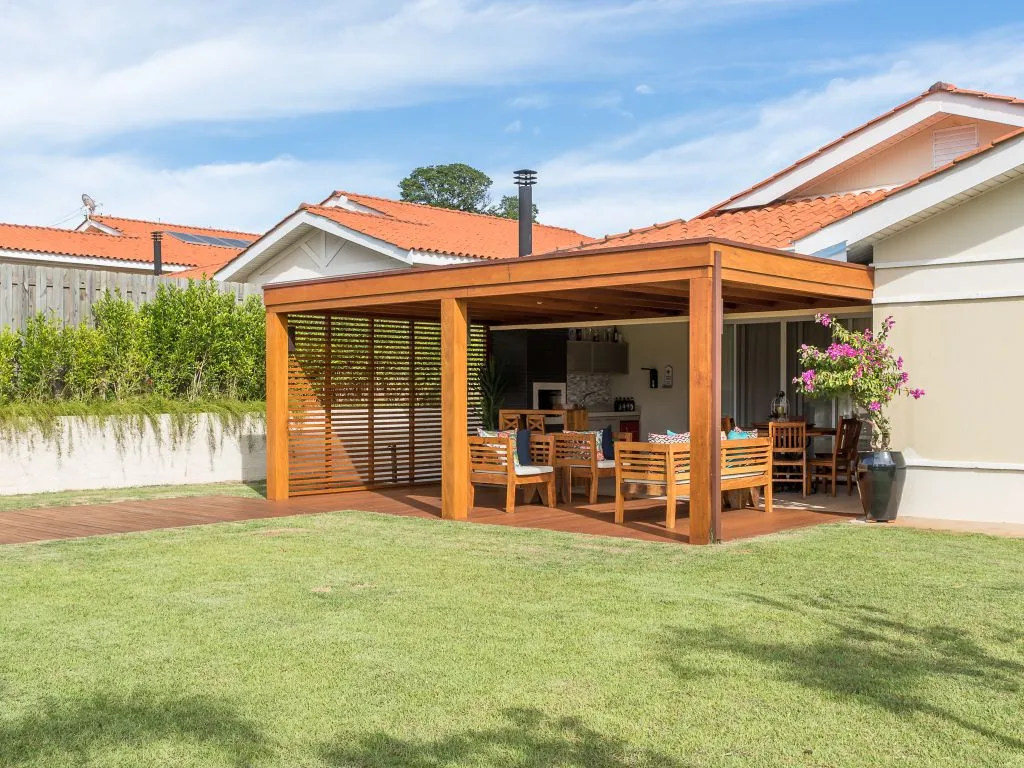 Pátio de uma casa com pergolado de madeira e móveis de exterior em madeira, visto de um amplo gramado. Uma parede de ripas de madeira oferece privacidade.